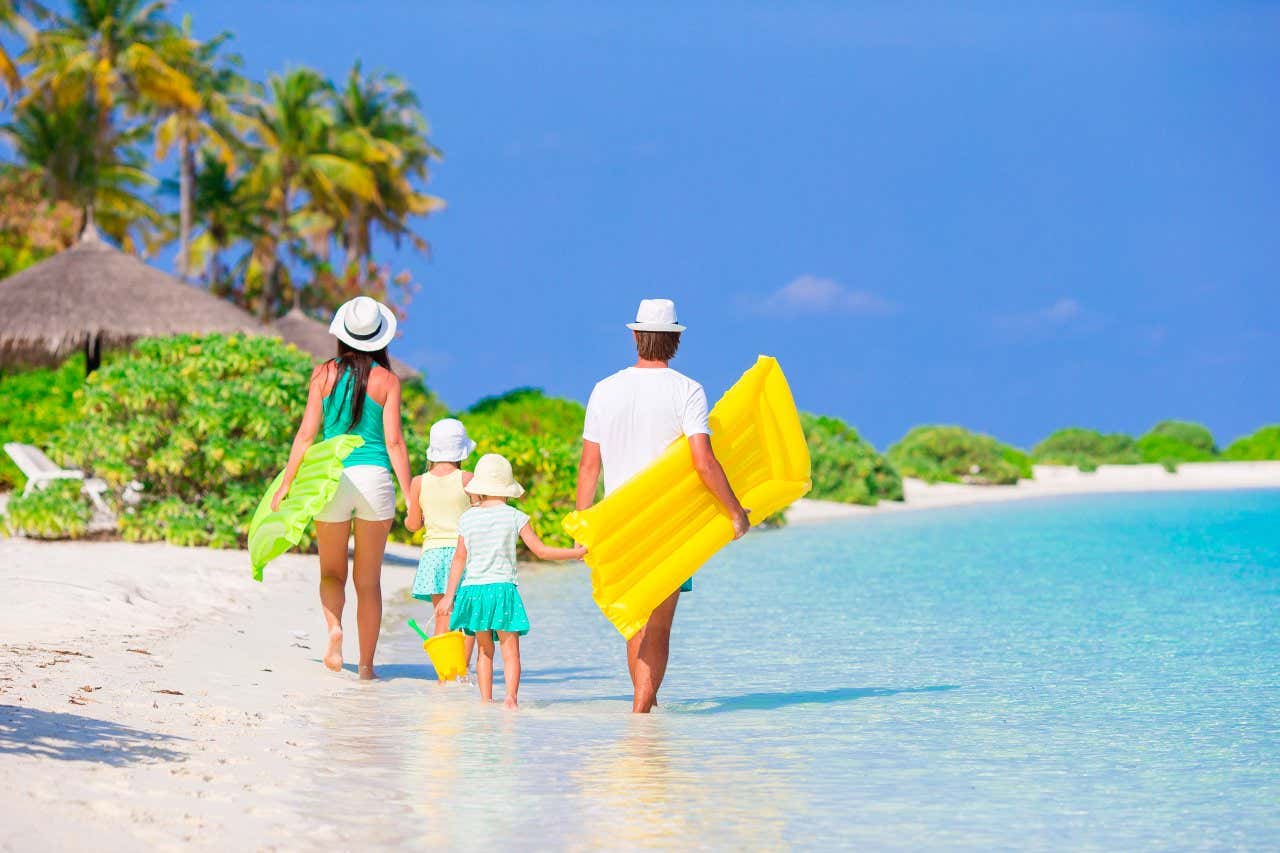Two adults and two children holding hands as they walk on a white sandy beach by the clear sea holding floats on their family vacation