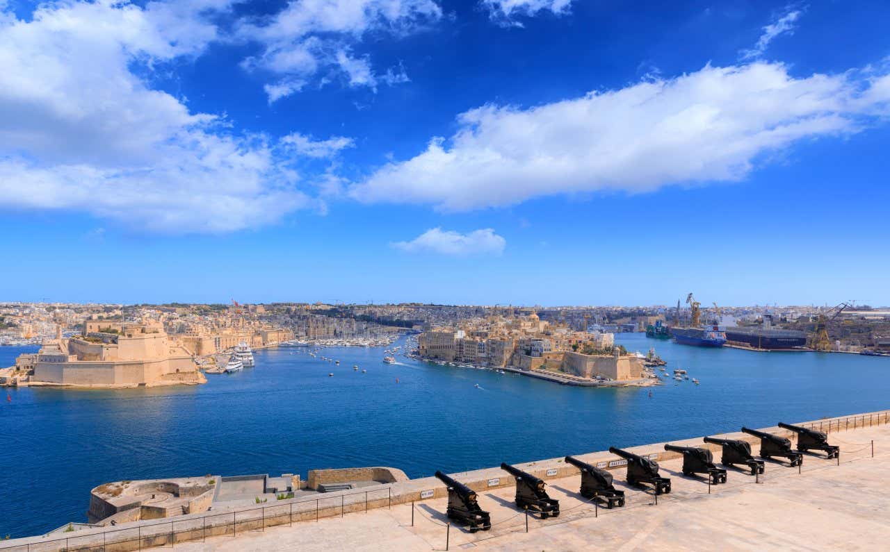 Una batteria di cannoni in fila affacciano sul golfo della capitale maltese, con sullo sfondo le Tre Città con alte mura e fortezze