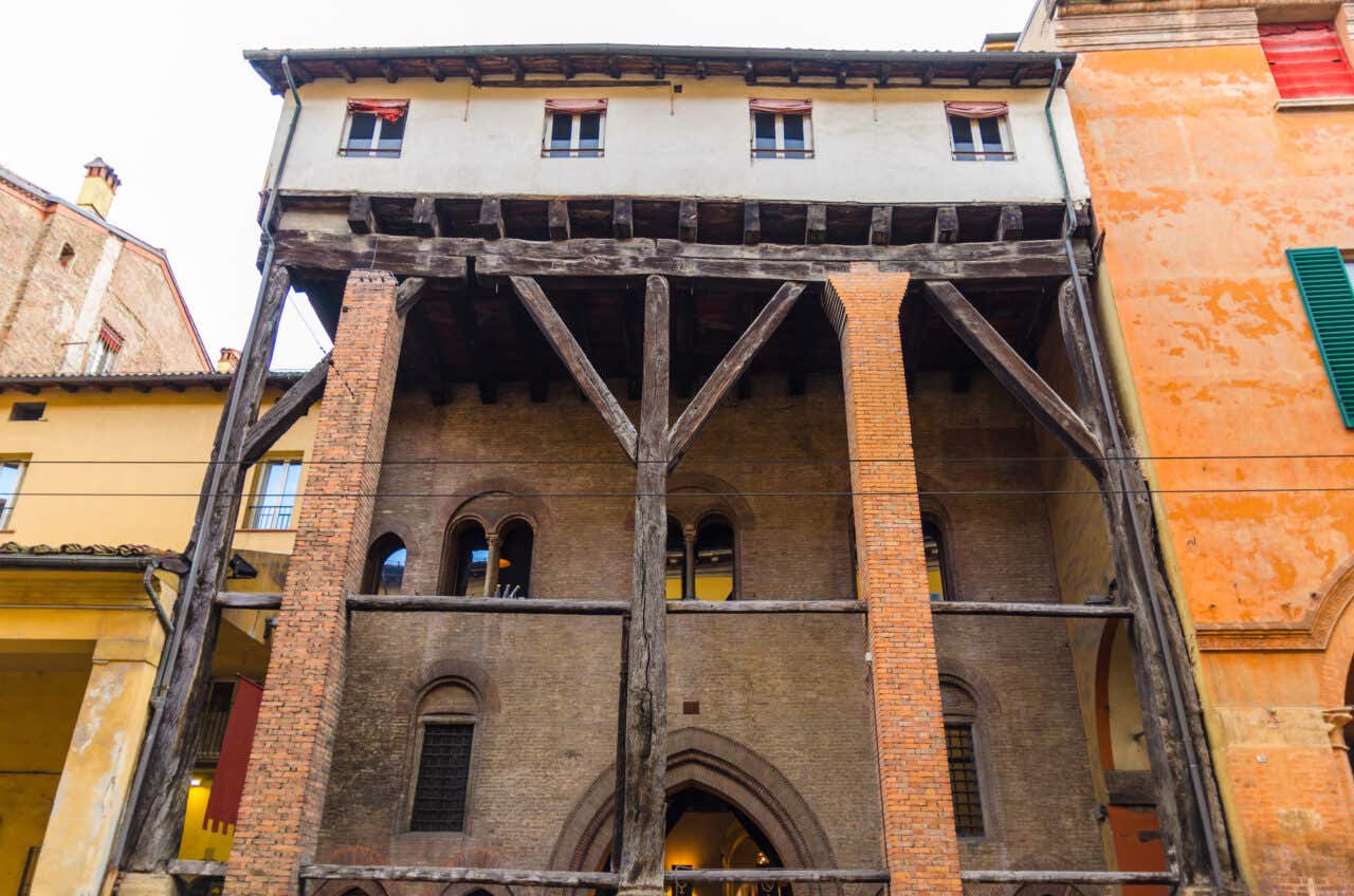 Edificio medievale nel centro storico di Bologna con facciata in mattoni e pietra, archi romanici e struttura lignea sporgente che crea un portico coperto, affiancato da edifici colorati gialli e arancioni