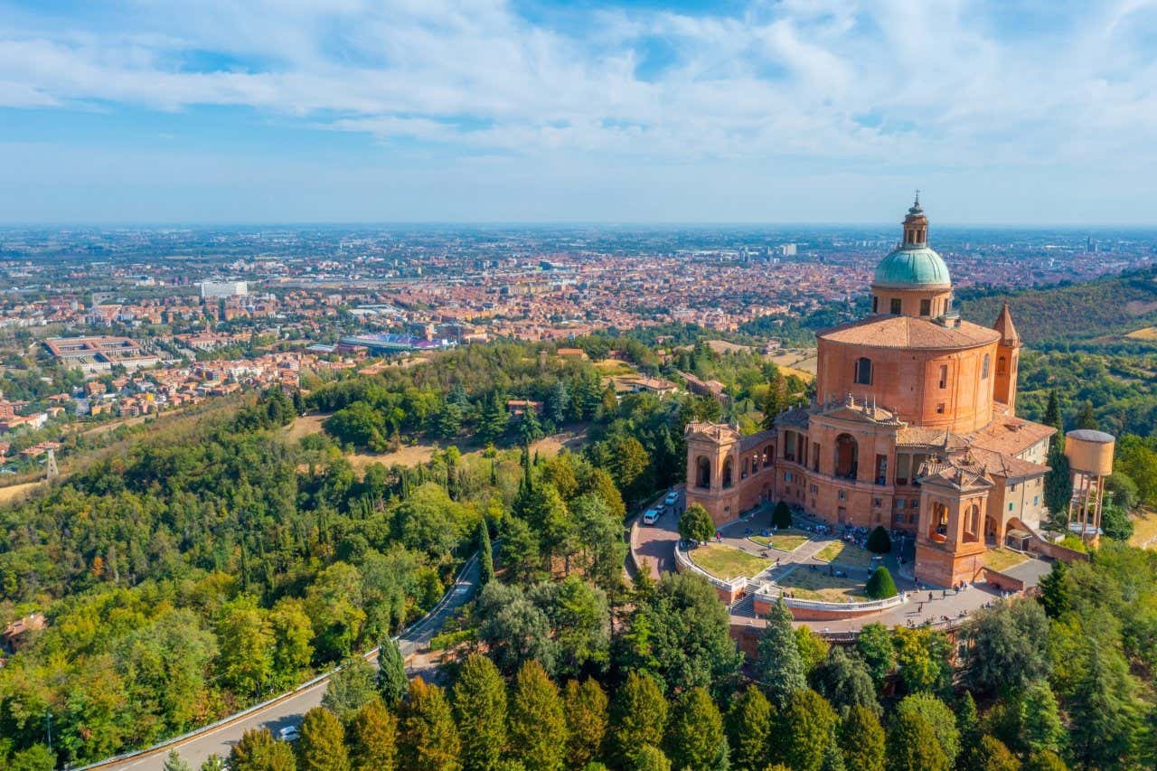 Vista aérea del Santuario de San Luca en Bolonia, rodeado de exuberante vegetación verde y con la ciudad al fondo, bajo un cielo azul despejado