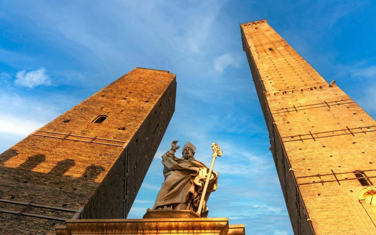 Las dos torres Garisenda y Asinelli vistas desde abajo con la estatua de un obispo en el centro. La torre Asinelli, a la derecha, es claramente más alta que la otra