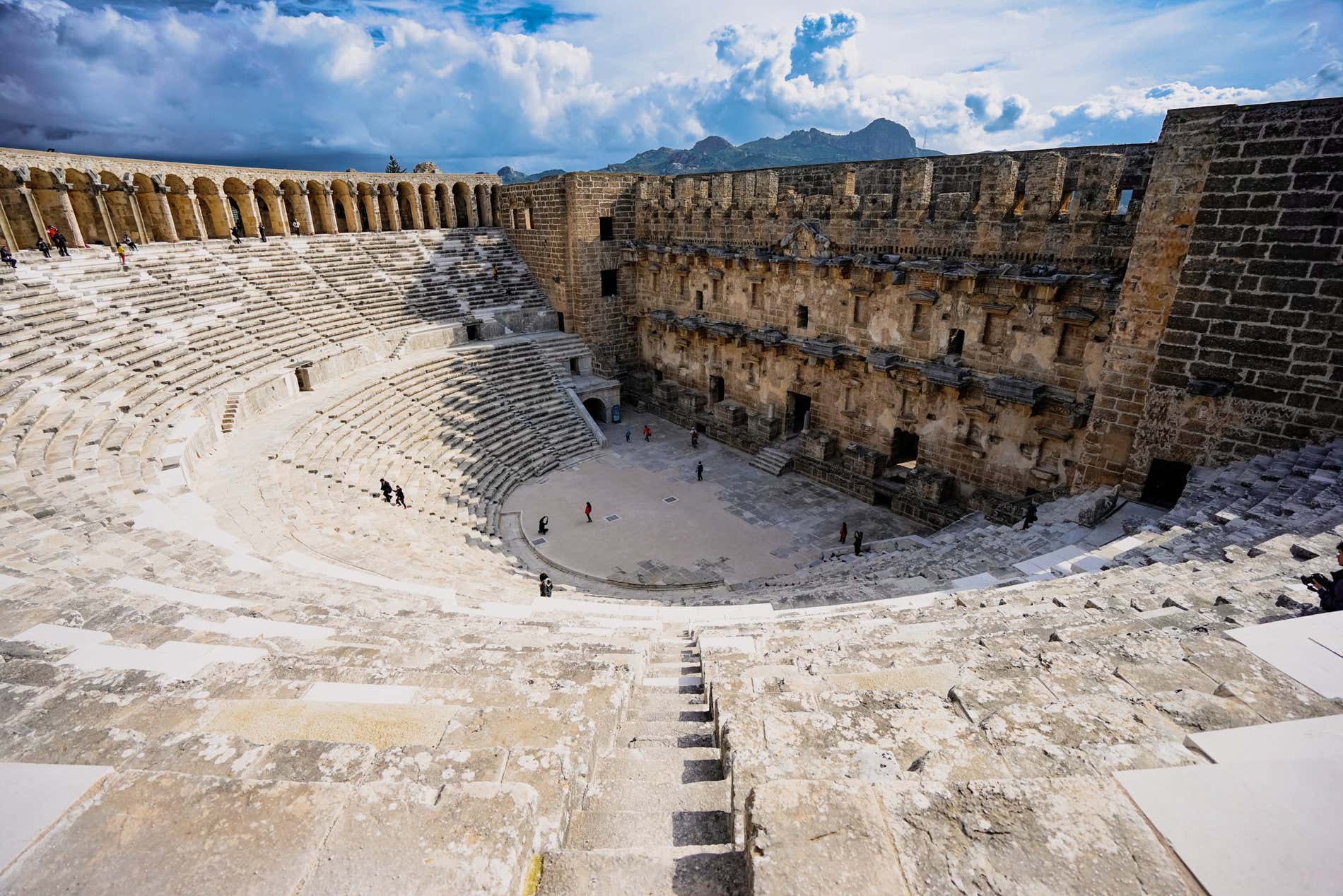 Il Teatro di Aspendos visto da uno degli scalini più in alto, con i turisti che passeggiano al suo interno e il panorama sullo sfondo.