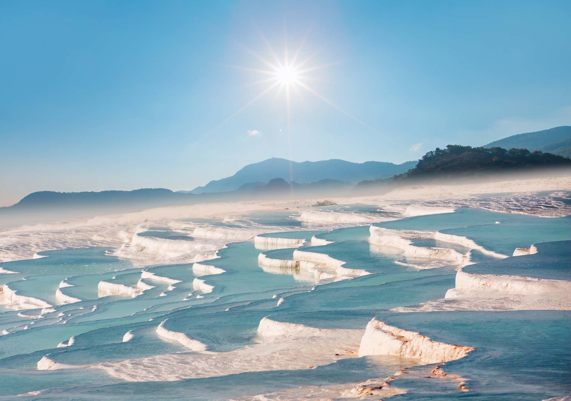 Pamukkale, il Castello di Cotone, con una leggera foschia che aleggia sul terreno e un cielo limpido con il sole sullo sfondo.