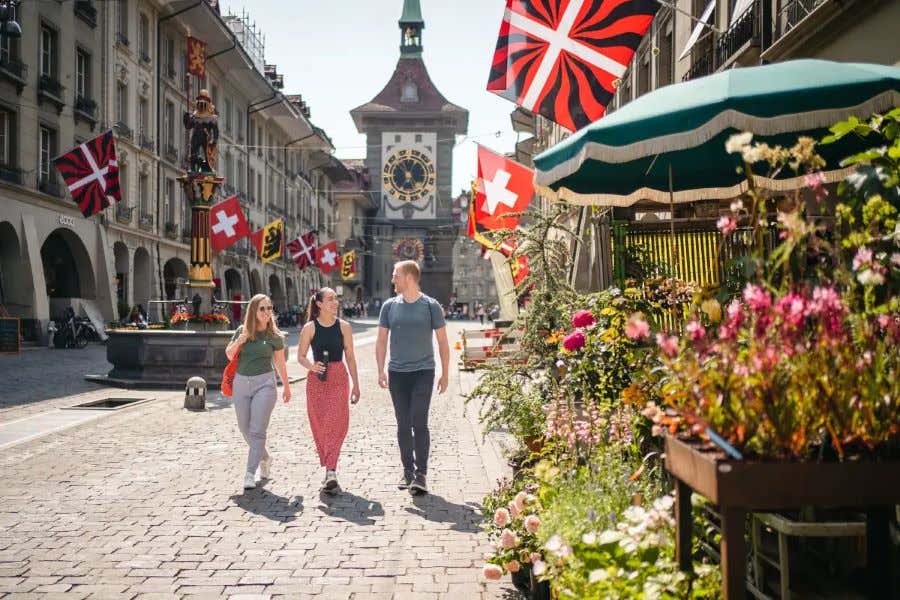 Varias personas caminan por una calle empedrada, con una torre con reloj de fondo