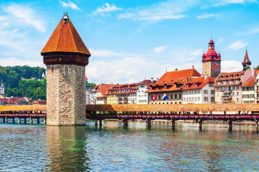 Puente de madera a orillas de un lago, con los campanarios de una ciudad de fondo