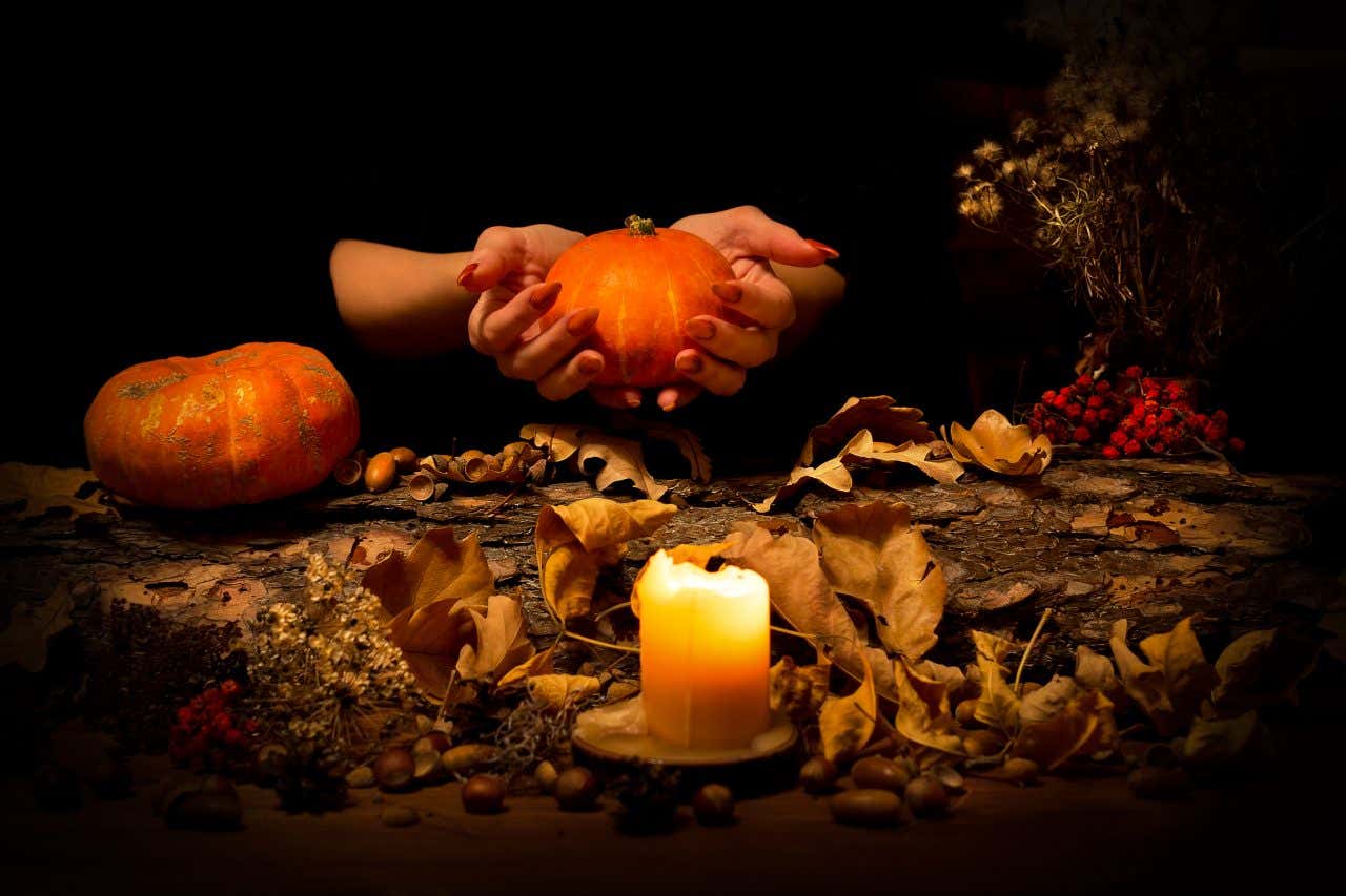 A pair of hands holding up a pumpkin over a leafy ground, with a candle lit in the foreground.