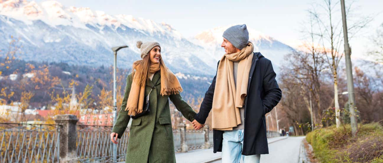 Una pareja de enamorados paseando con ropa de invierno frente a un pueblo con vistas a unas montañas nevadas