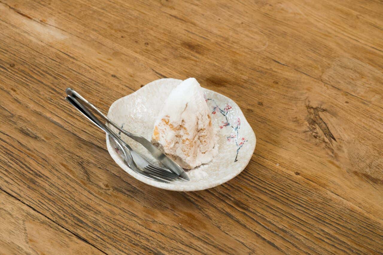 Un piccolo dolce a forma trapezoidale ricoperto di zucchero a velo e servito su un piattino di ceramica al centro di una tavola in legno