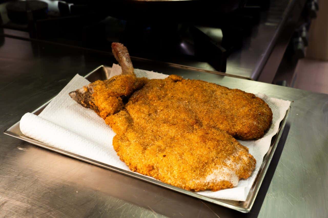 Una cotoletta di carne di vitello impanata e fritta poggiata su una teglia su un ripiano in metallo all'interno della cucina di un ristorante