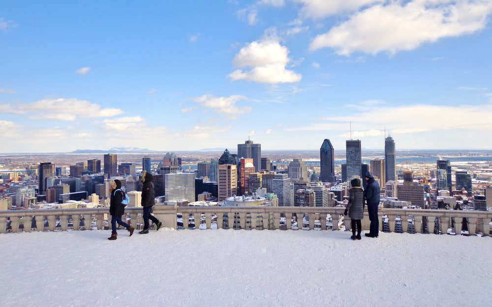 Una serie di persone in abiti invernali ammira i grattacieli di Montréal dall'alto di un belvedere in inverno, con la neve a terra e sui palazzi