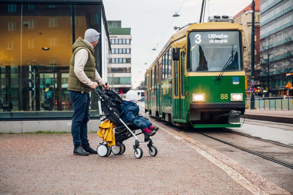 Un uomo in vestiti invernali con un bimbo in passeggino, attendono il passaggio di un piccolo tram urbano tra i palazzi moderni del centro di Helsinki
