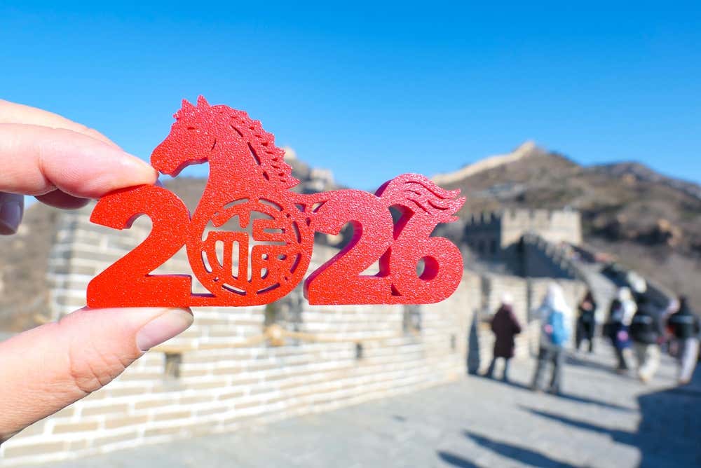 Mano che regge una decorazione rossa a forma di "2026" con un cavallo stilizzato e il carattere cinese per fortuna e prosperità, fotografata davanti alla Grande Muraglia Cinese sotto un cielo azzurro. Sullo sfondo si vedono alcuni visitatori e le torri di avvistamento della muraglia. La decorazione celebra il Capodanno Cinese 2026, Anno del Cavallo