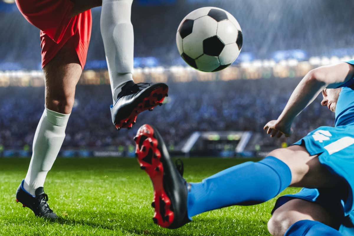 Escena de un partido de fútbol en el que se ve un balón y dos jugadores, uno con medias rojas y otro con medias azules