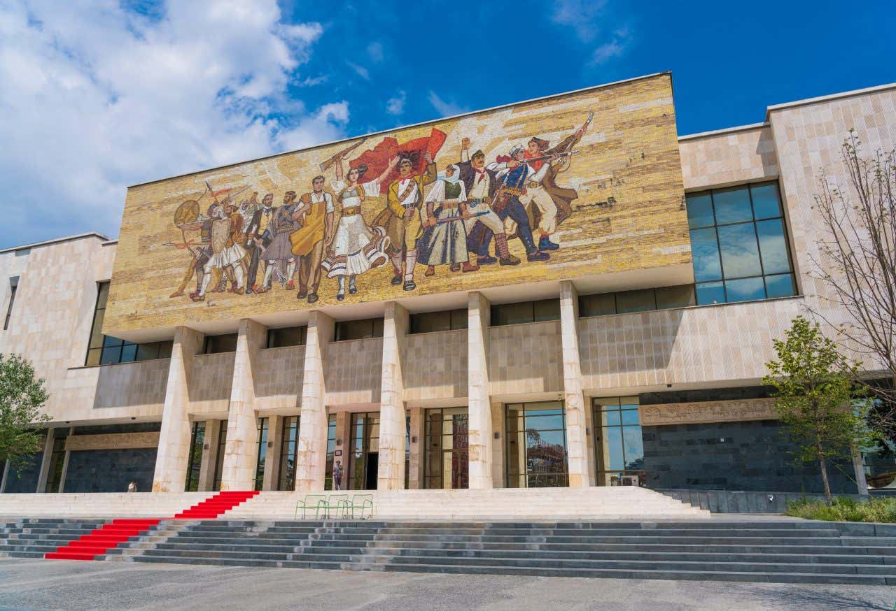 Facciata di un edificio bianco sormontata da una grande pannello di pietra con un mosaico policromo raffigurante il popolo albanese che marcia con la bandiera dell'Albania, un'aquila nera su sfondo rosso