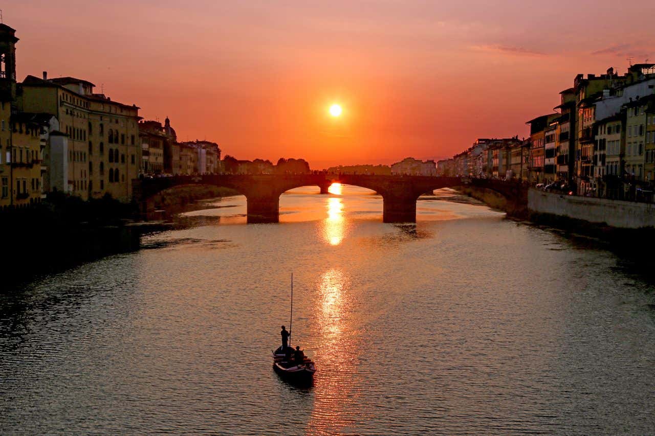 The Arno river with a traditional gondola boat sailing and a bridge in the distance at sunset