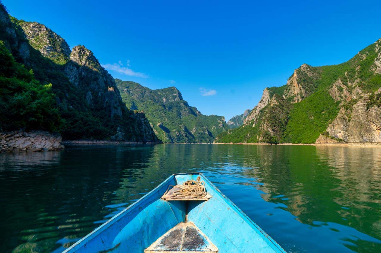 Foto scattata da una piccola imbarcazione di cui si vede la prora, naviga lungo un lago circondato da montagne in un giorno di sole senza nuvole