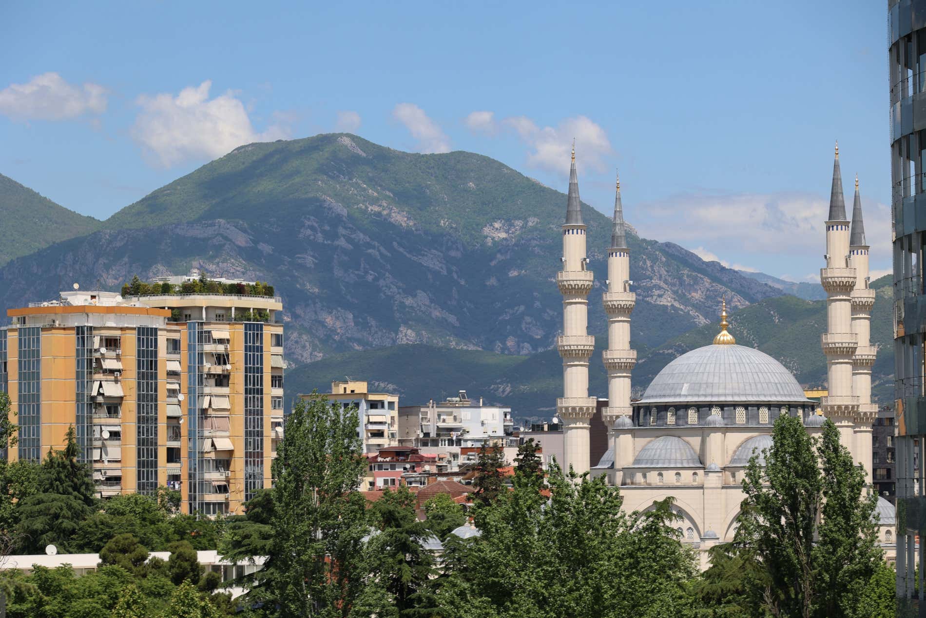 Foto panoramica di una moschea di Tirana accanto a un palazzo e un monte ricoperto di boschi sullo sfondo