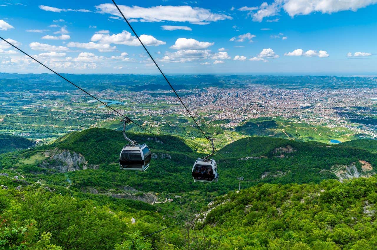 Due cabine appese a due fili che salgono o scendono dalla cima di un monte, alle loro spalle il profilo della città di Tirana