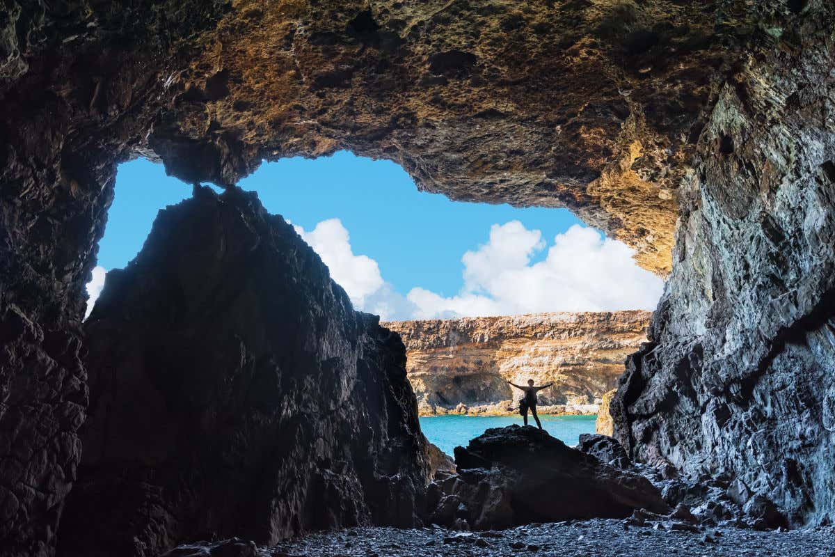 Una persona con los brazos estirados subida en una roca en mitad de una enorme cueva con vistas al mar