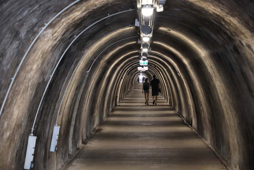 Foto di un tunnel illuminato da luci elettriche e una porta in fondo che dà all'esterno da cui entra molta luce. In mezzo al corridoio, due persone camminano di spalle verso l'uscita