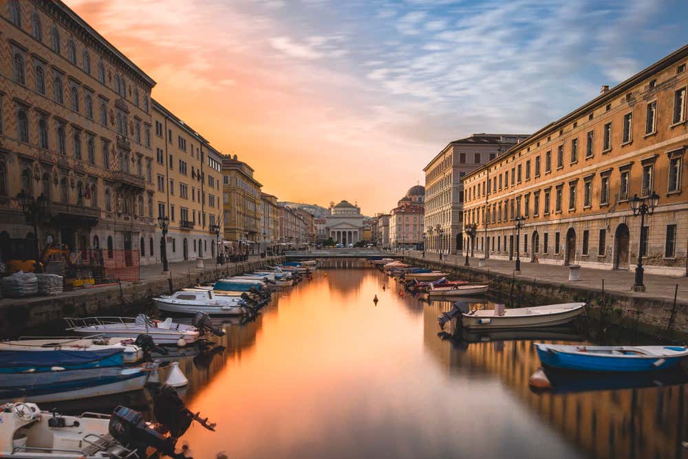 Un canale d'acqua i mezzo ai palazzi del centro storico, interrotto sul fondo da una chiesa neoclassica. L'acqua del canale riflette la luce arancione del cielo.