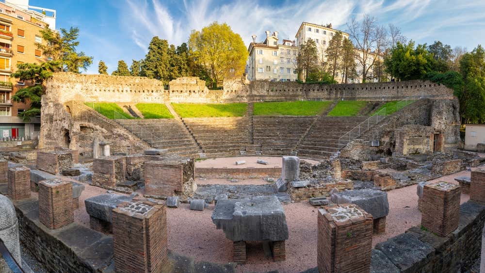 Una scalinata a semicerchio con i resti di colonne in mezzo ai palazzi e alberi del centro storico di Trieste