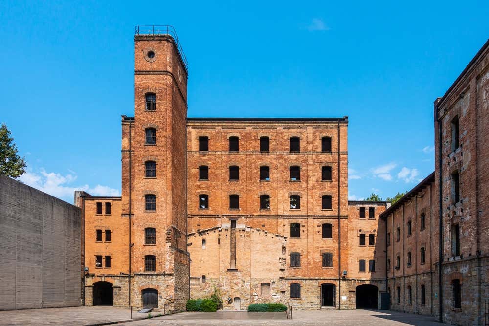 Una grande edificio in mattoni rossi fotografato dal cortile interno in cemento. Sulla facciata si aprono tante finestrelle nere, segnalando la presenza di sei piani totali