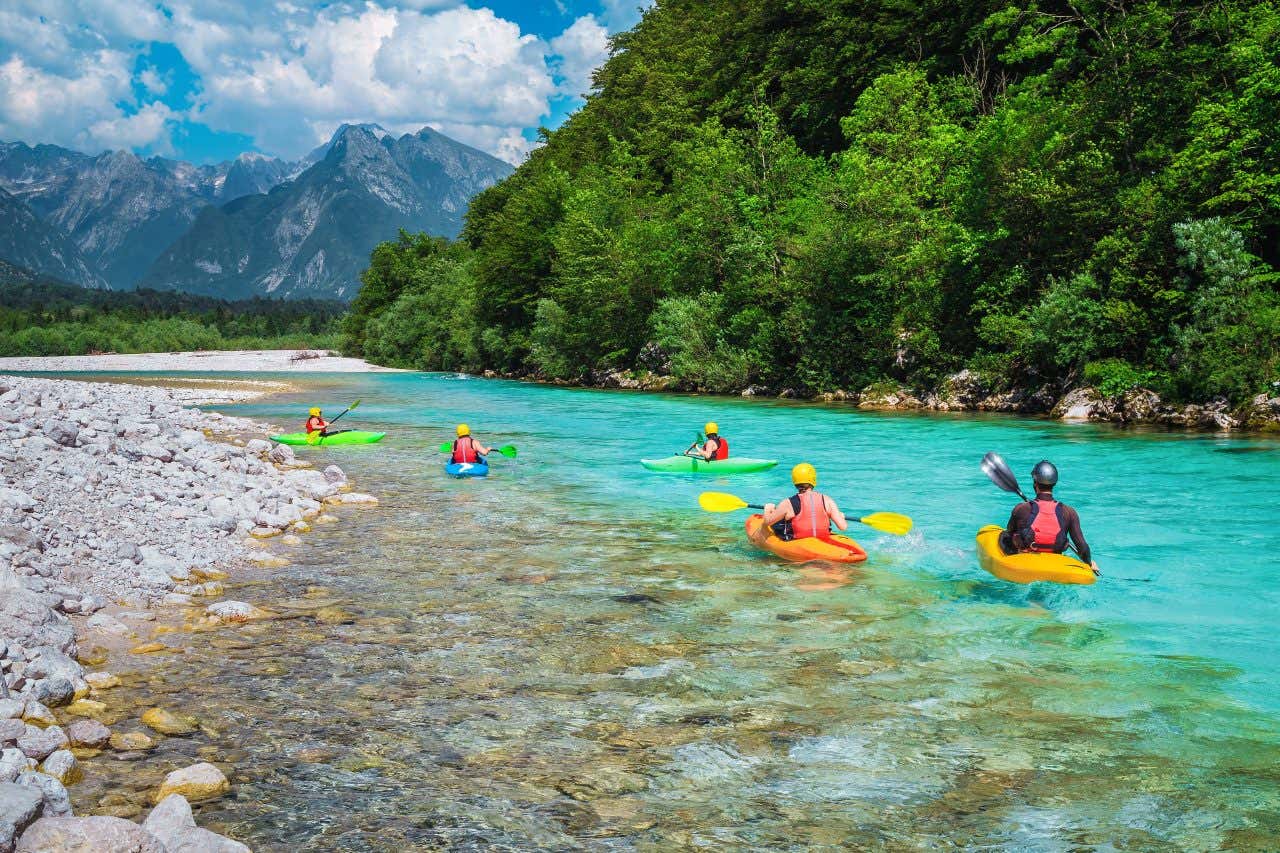 Cinque persone in kayak riprese di spalle navigano lungo un fiume, accanto a un bosco rigoglioso e con una catena montuosa sullo sfondo