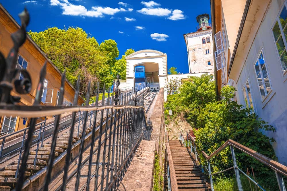 In mezzo ad alcuni palazzi, fotografata dal basso, c'è una rampa in pietra con binari, sormontata da una piccola stazione da cui parte la cabina della funicolare di colore blu
