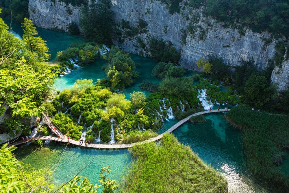 Vista aerea di alcuni escursionisti che attraversa una passerella di legno costruita sopra un lago turchese nei pressi di alcune cascatelle all'interno di una gola carsica