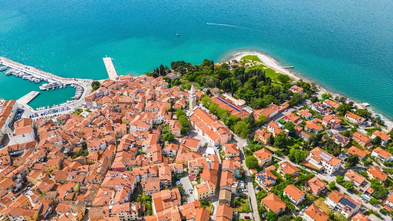 Vista del centro di Izola dall'alto. Si vedono i tetti arancioni delle case e un piccolo porticciolo che si estende sul mare turchese dell'Adriatico
