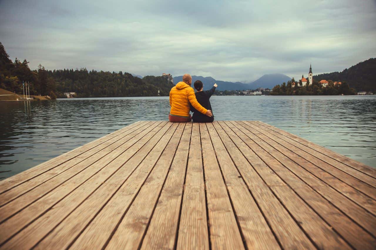 Una coppia di spalle seduta su un molo osserva il Lago di Bled e la sua isola