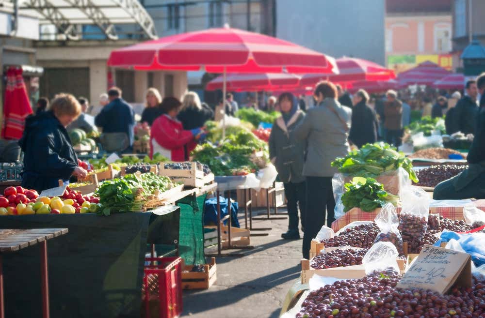 Un mercato all'aperto nel centro di Zagabria pieno di bancarelle che vendono frutta e verdura e diverse persone, tra commercianti e clienti, che passeggiano e lavorano