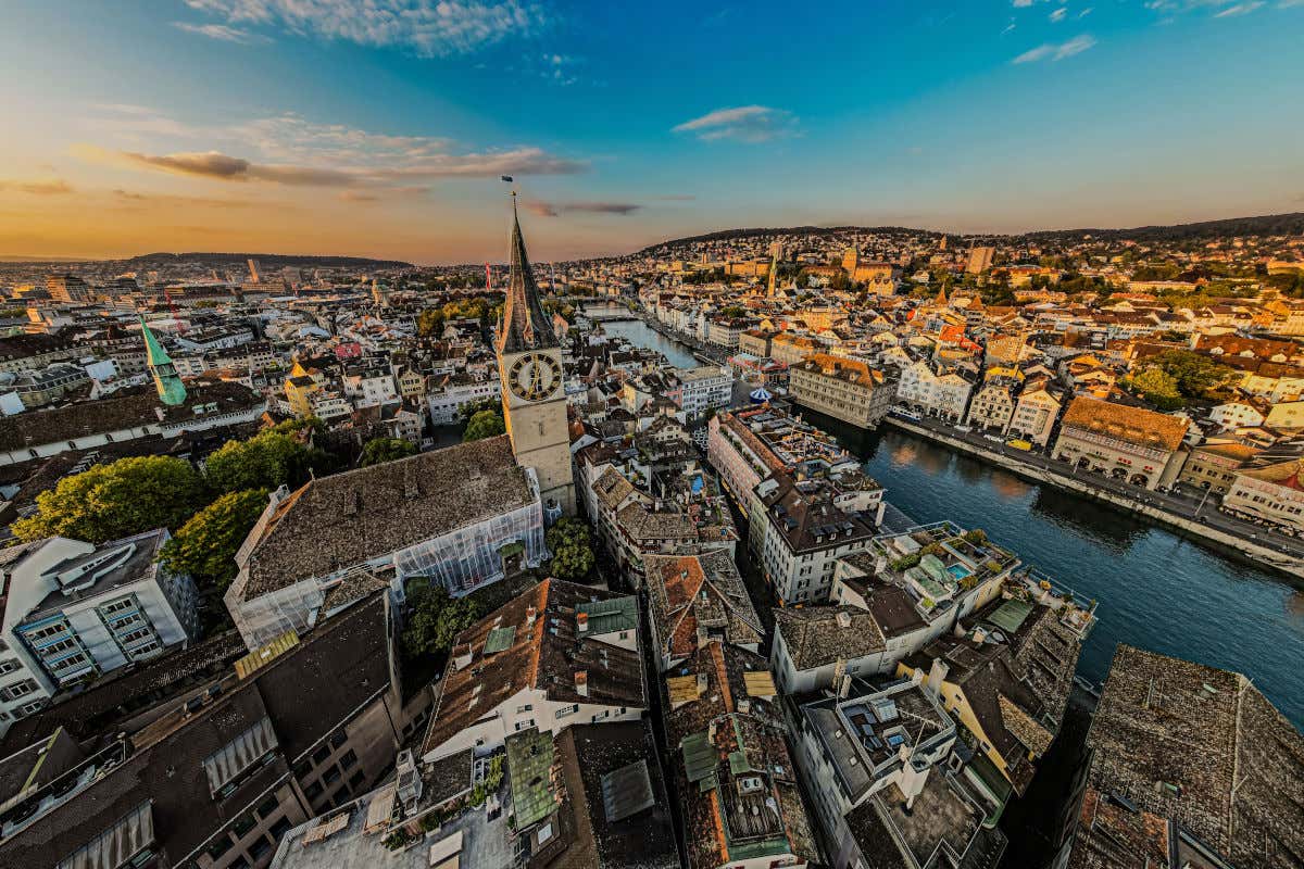 Vista aérea de Zúrich con varios edificios y un canal de agua dividiendo la ciudad, destacando una iglesia con torre del reloj