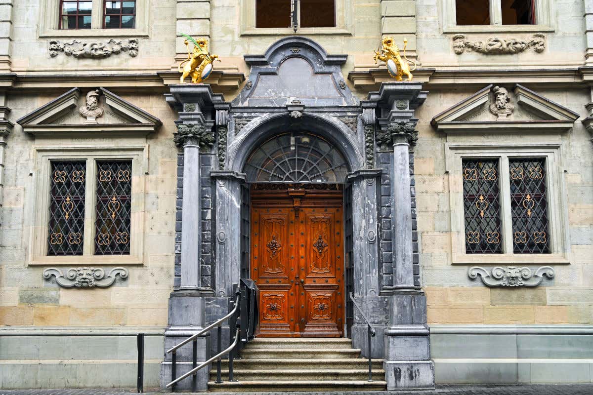 Un elegante pórtico de un Ayuntamiento con puerta de madera, escaleras de piedra y dos esculturas de leones dorados sujetando un escudo