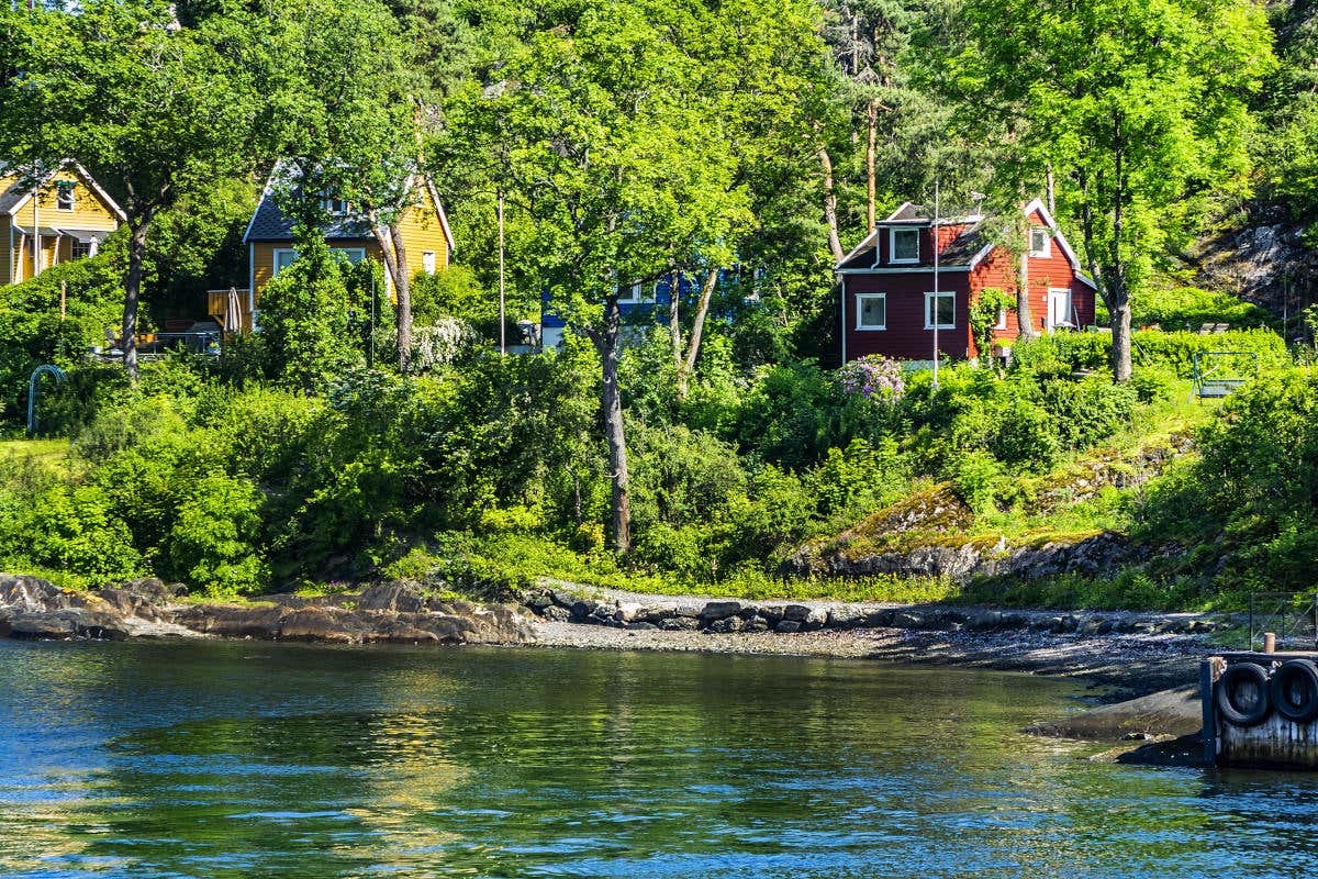 Varias casas de madera, de colores rojizos y amarillos, rodeadas de vegetación frente a las aguas esmeraldas del fiordo de Oslo