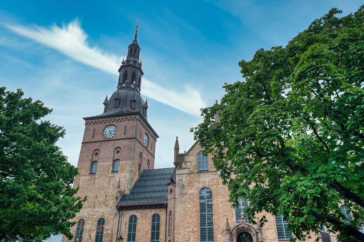 Una catedral de fachada de ladrillo y con una torre de reloj frente a un conjunto de árboles