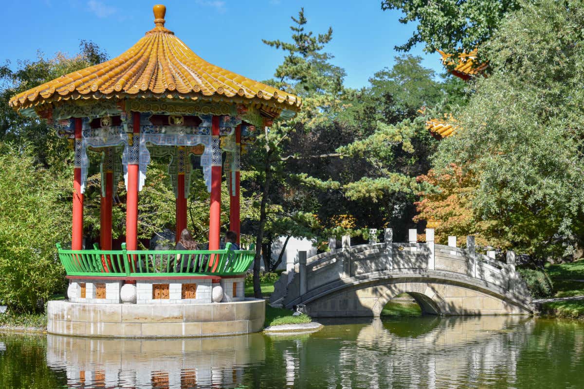 Un estanque con un pequeño puente de piedra y un templete de inspiración asiática rodeado de árboles