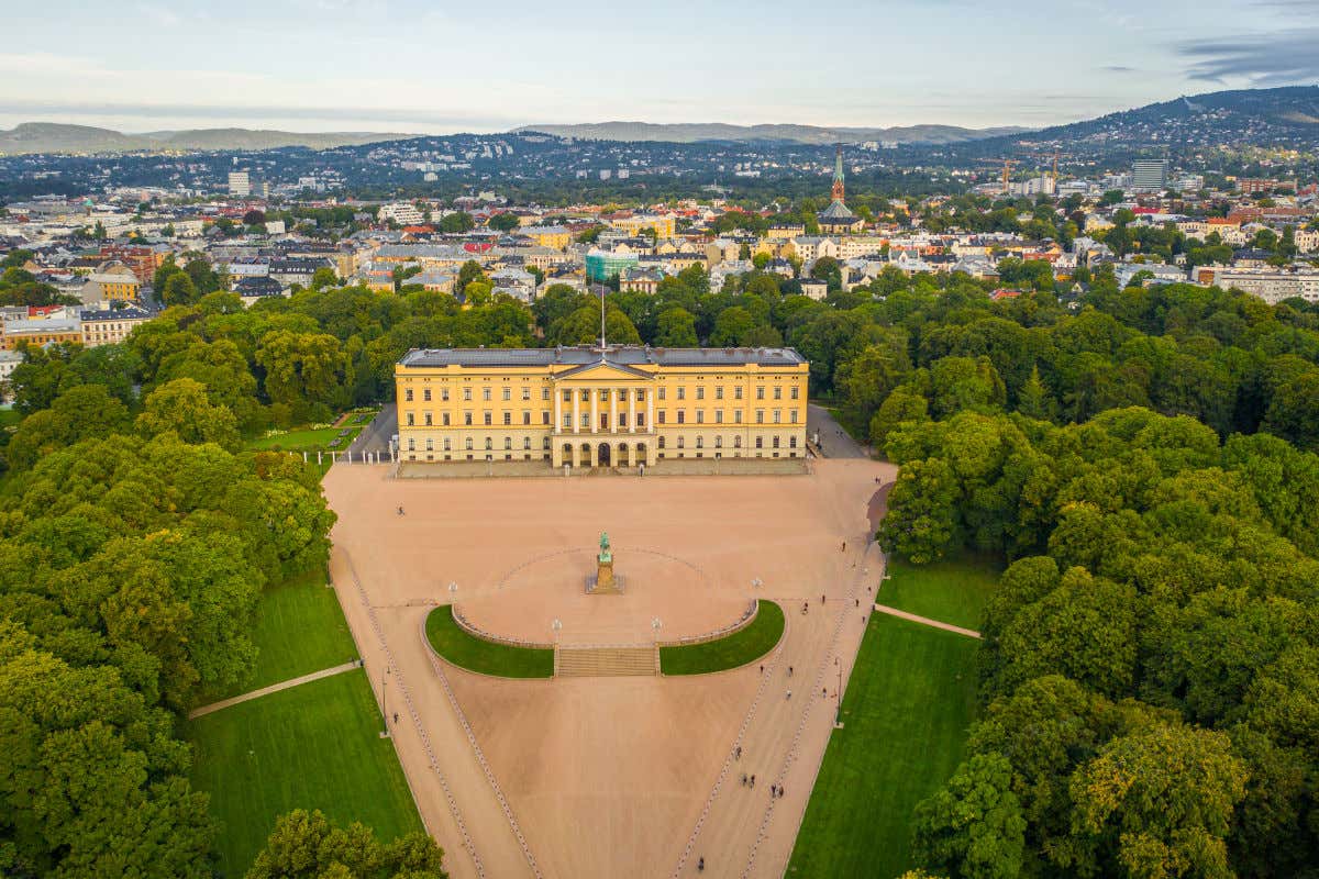 Vista aérea del Palacio Real de Oslo, un edificio de color amarillo en mitad de una explanada rodeada de árboles y con una escultura frente a su pórtico
