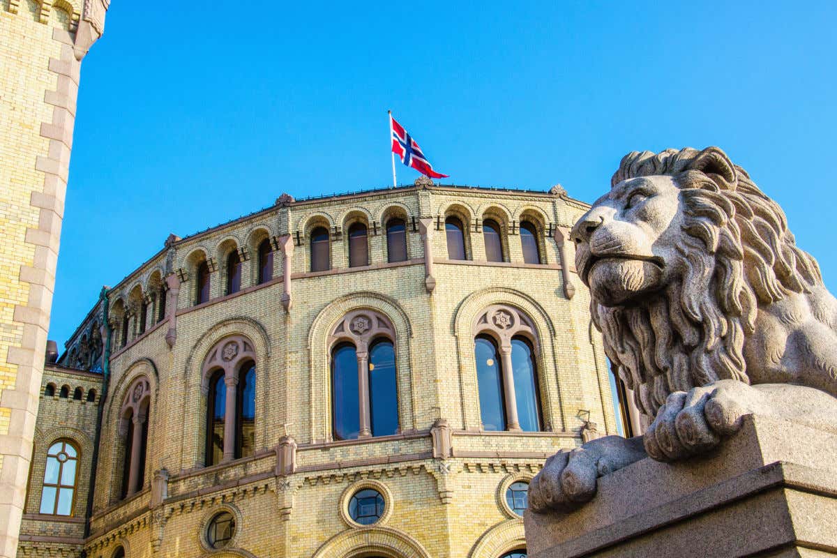 Escultura de un león de piedra frente a un edificio de escasa decoración pero con una bandera de Noruega
