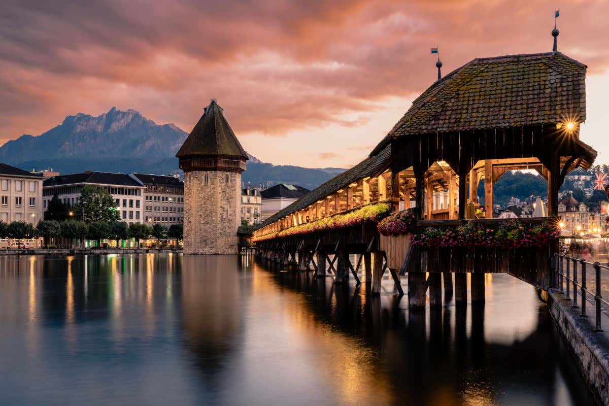 Un puente de madera de origen medieval iluminado por la noche y junto a una torre de piedra en la ciudad de Lucerna