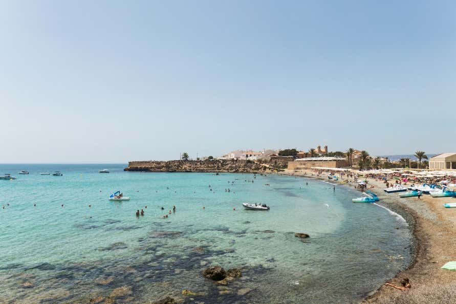 A view of a beach on the Tabarca Island, with boats anchored near the shore, under a clear blue sky.