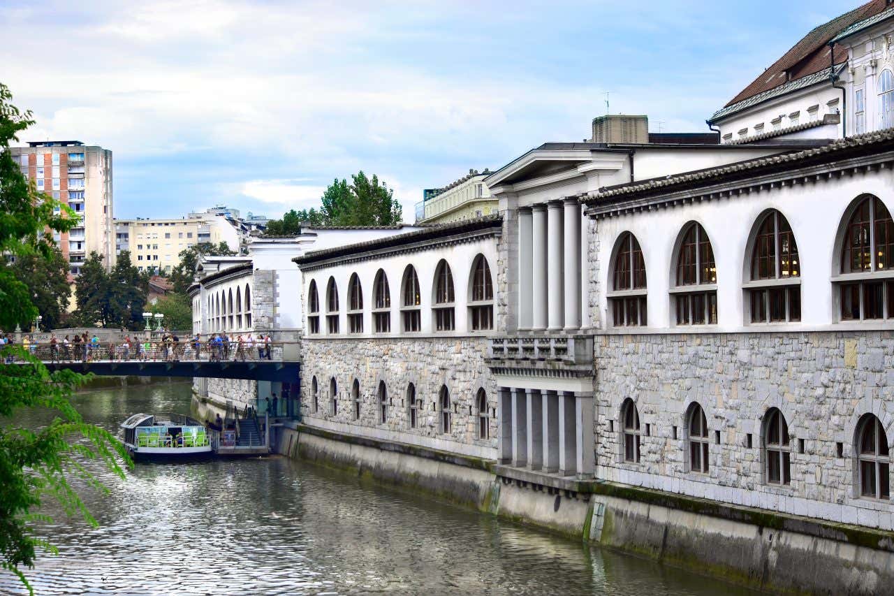 Un edificio costruito lungo i bordi del fiume. Si tratta del mercato coperto di Lubiana, costruito su due piani e separato al centro da una struttura che ricorda un tempio greco
