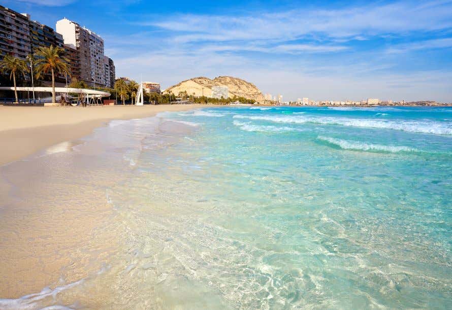 A view of El Postiguet Beach, with turquoise water and golden sand and a rock formation in the distance under a blue sky.