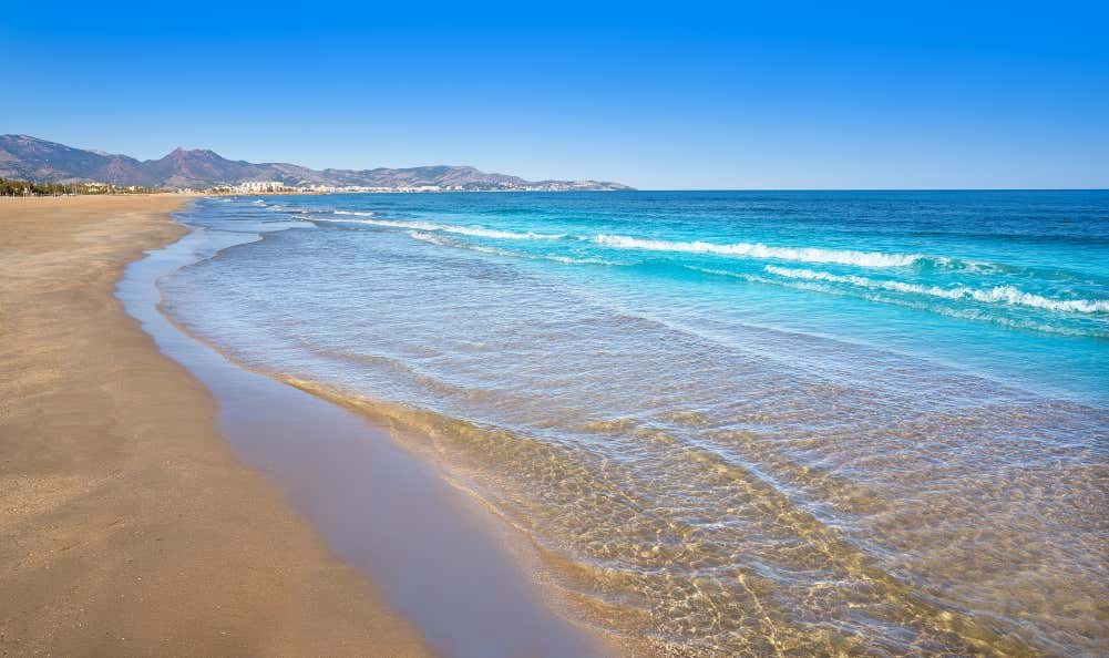 A view of El Gurugú Beach, a sandy beach with turquoise water and calm waves crashing on the shore and mountains beyond, under a clear blue sky.