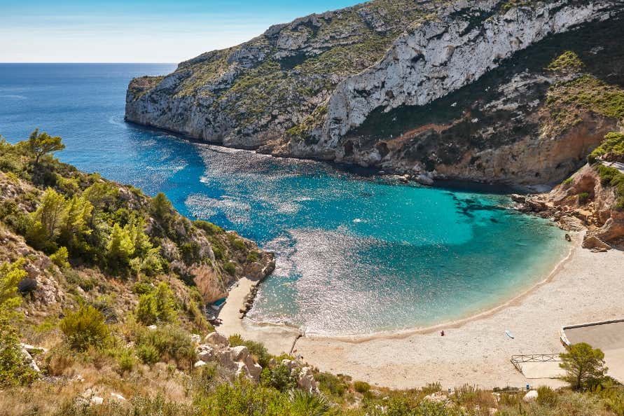 A view of a beach in a cove, surrounded by mountains, with turquoise waters and golden sand.