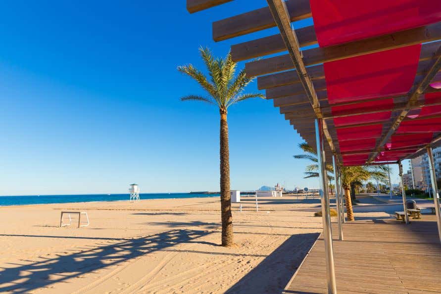 A view of the Gandia Beach, a sandy beach with palm trees and a white watch tower under a clear blue sky.