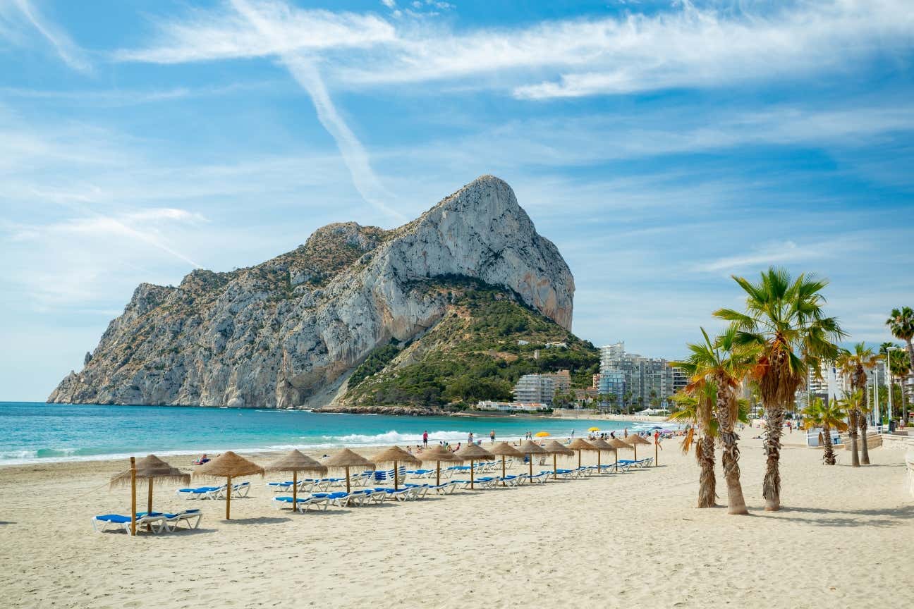 A view of a sandy beach with sun umbrellas and palm trees, and a rock formation seen across it.