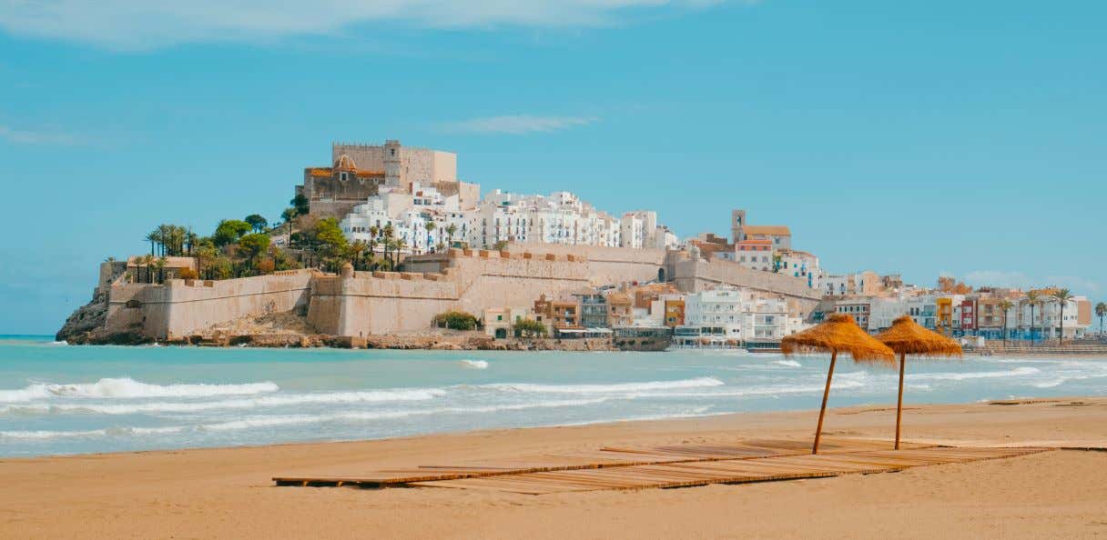 A view of Peñíscola Castle from El Nord Beach in Castellón, a sandy beach with two straw umbrellas on it.