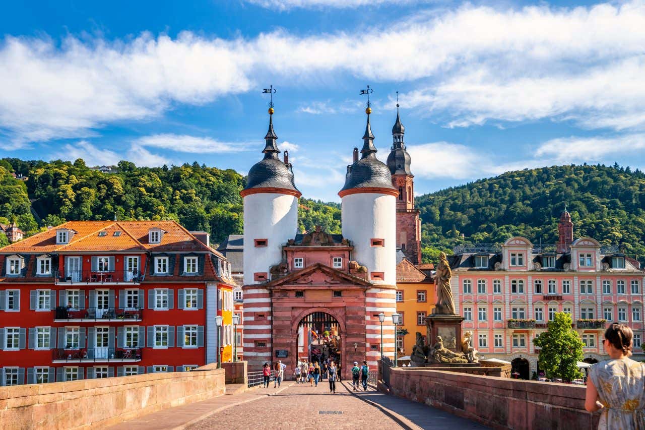 View looking down the Old Bridge in Heidelberg, the city gate with two towers rising above colorful houses on either side on a sunny day